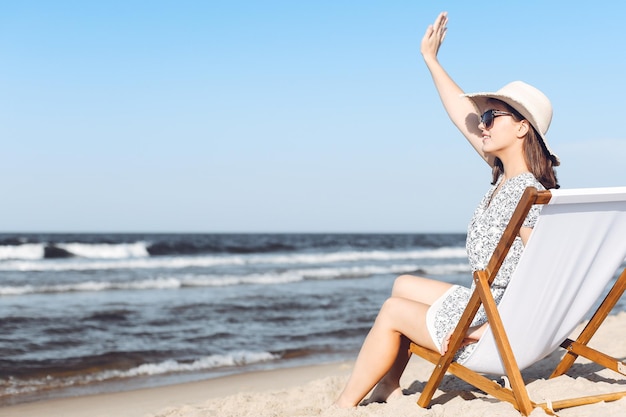 Mujer morena feliz sentada en una tumbona de madera en la playa del océano mientras saluda y saluda a alguien con la mano.