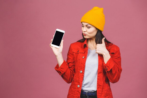 Mujer morena feliz sorprendida en la pantalla casual en blanco que muestra smartphone. Pulgares hacia arriba.