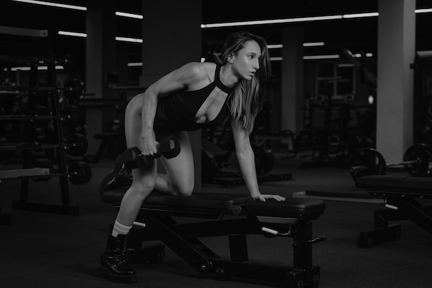 Una mujer morena en forma está haciendo una fila con mancuernas de un solo brazo con la rodilla en el banco. Una chica musculosa viste un stringbody negro y botas durante un entrenamiento de espalda en un gimnasio.