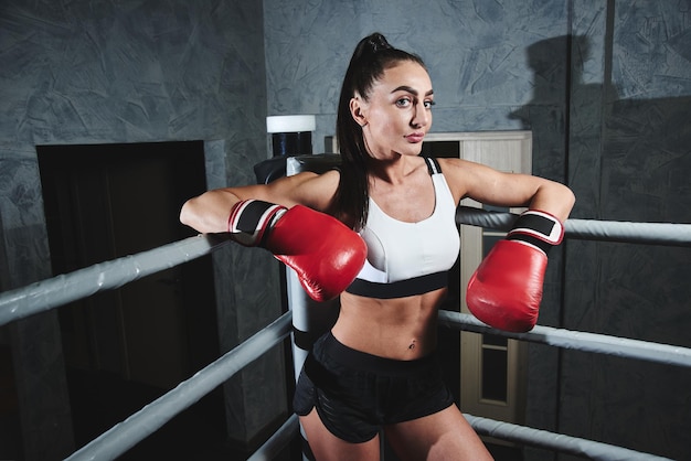 Una mujer morena fuerte y sexy con top de ropa deportiva y pantalones cortos con guantes de boxeo en el ring mira a la cámara