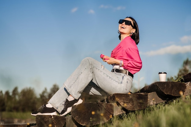 Mujer morena con gafas de sol y camisa rosa sentada y relajada en el parque sobre el fondo del cielo azul