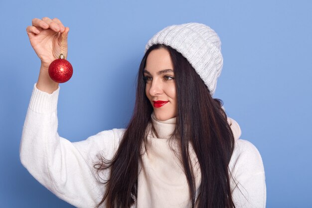 Mujer morena con gorra blanca y suéter, sosteniendo la bola roja de Navidad en las manos y mirándola