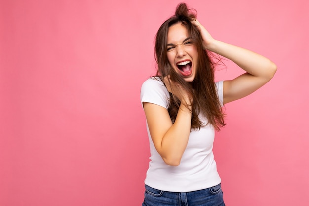 Mujer morena hermosa joven. Señora de moda en camiseta blanca casual de verano para maqueta. La mujer positiva muestra emociones faciales sinceras. Modelo divertido aislado sobre fondo rosa con espacio libre.