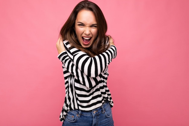 Mujer morena hermosa joven. Señora de moda en manga larga a rayas casual. La mujer positiva muestra emociones faciales sinceras. Modelo divertido aislado sobre fondo rosa con espacio libre.