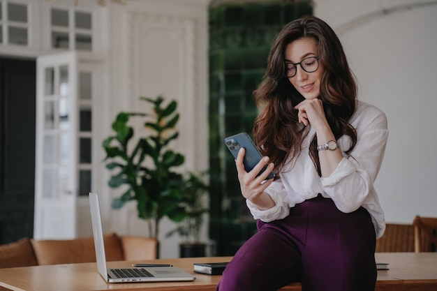 Mujer morena hermosa y pensativa con gafas, camisa blanca y pantalones violetas, sentada en la mesa sosteniendo el teléfono desplazándose por Internet Empresaria complacida hablando con su hija a través de una videollamada Gente de negocios