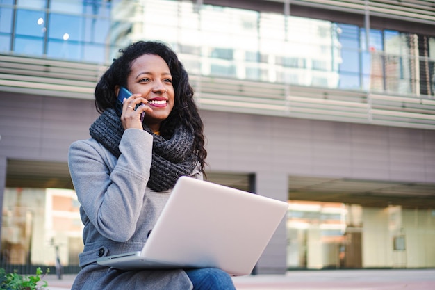 Mujer morena impresionante sonriente con ropa informal sentada al aire libre con una computadora portátil mientras habla por teléfono inteligente Concepto de negocio