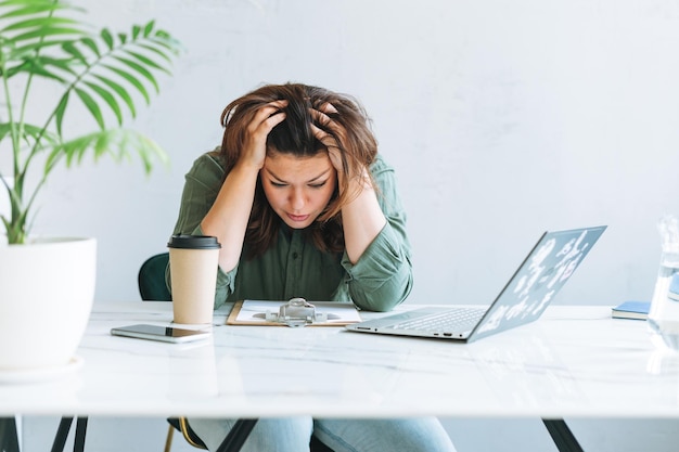 Mujer morena infeliz de pensamiento joven de talla grande que trabaja en una computadora portátil en la mesa con una planta en la oficina moderna y luminosa