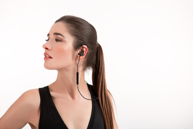 Mujer morena en jogging top negro escuchando música con auriculares posando en gris
