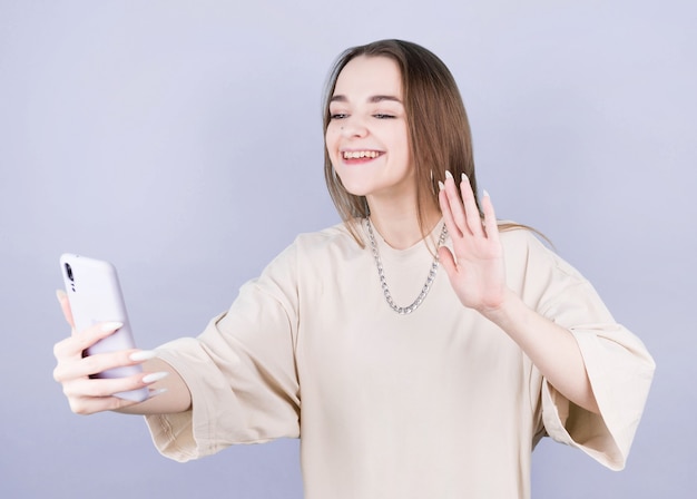 Mujer morena joven alegre emocionada en top sin mangas beige liso de pie haciendo selfie en teléfono móvil saludando con la mano saludo aislado en la pared de color púrpura, retrato