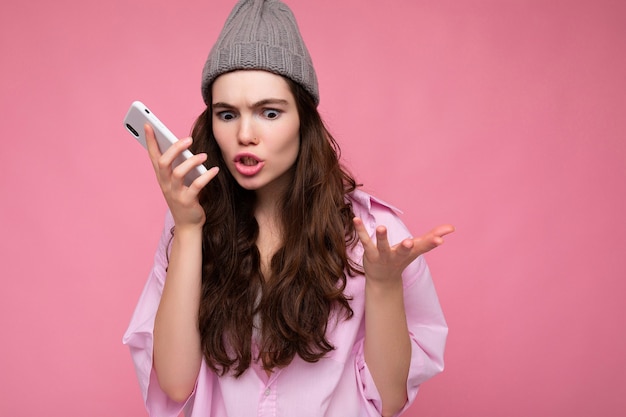 Mujer morena joven atractiva enojada emocional que lleva la camisa rosada elegante y el sombrero gris aislado sobre fondo rosado que sostiene en la mano y que usa el teléfono móvil que se comunica y graba el mensaje de voz.