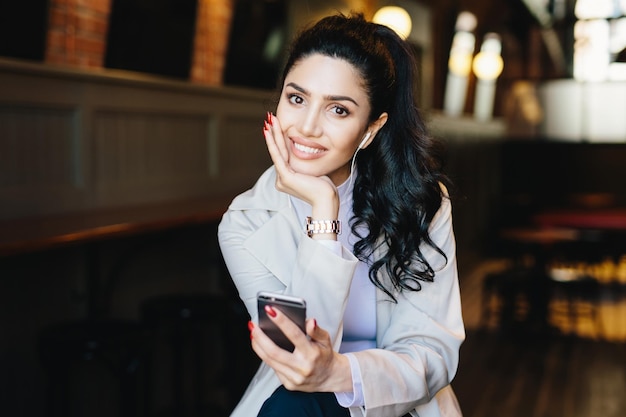 Mujer morena joven caucásica con cola de caballo larga con cara bonita y manos suaves con manicura roja escuchando su música favorita con auriculares y teléfono celular posando en el fondo del café