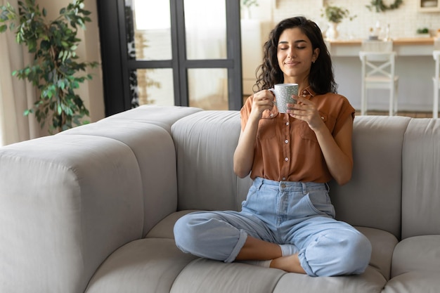 Mujer morena joven pacífica sentada en el sofá con los ojos cerrados bebiendo café caliente en casa copia