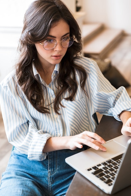 Mujer morena joven pensativa atractiva que estudia en el café en el interior, trabajando en la computadora portátil