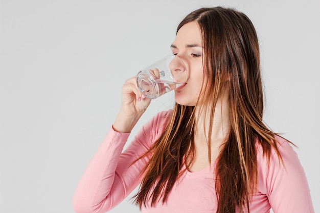 Mujer morena joven que bebe el agua pura aislada en fondo gris