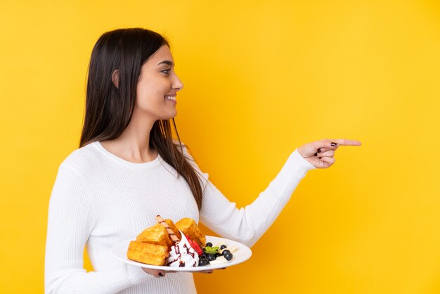 Mujer morena joven que sostiene las galletas sobre la pared aislada que señala al lado para presentar un producto