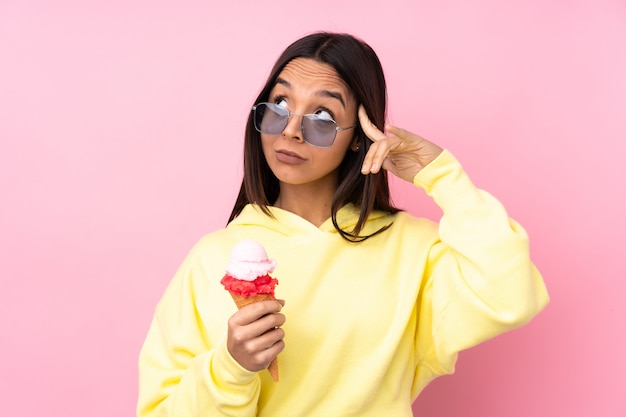 Mujer morena joven que sostiene un helado de cucurucho sobre la pared rosada aislada que tiene dudas y pensamiento