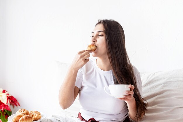 Mujer morena joven sentada en la cama con croissants comiendo