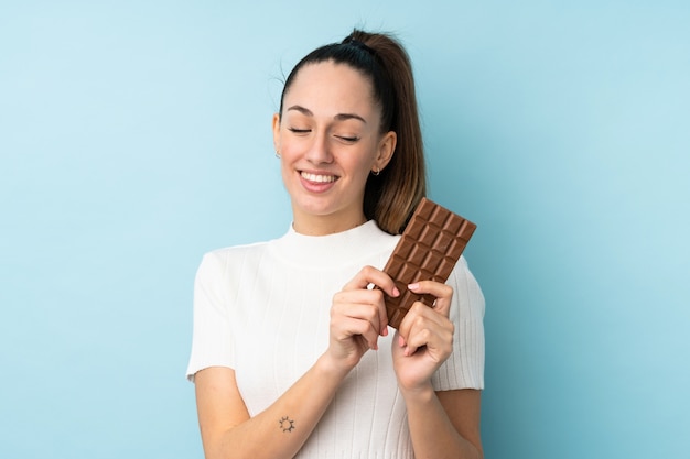 Mujer morena joven sobre la pared azul aislada que toma una tableta de chocolate y feliz