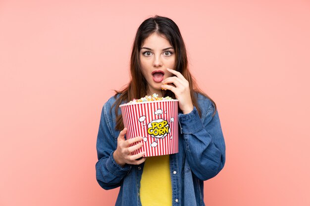 Mujer morena joven sobre la pared rosada aislada que sostiene un cubo grande de palomitas