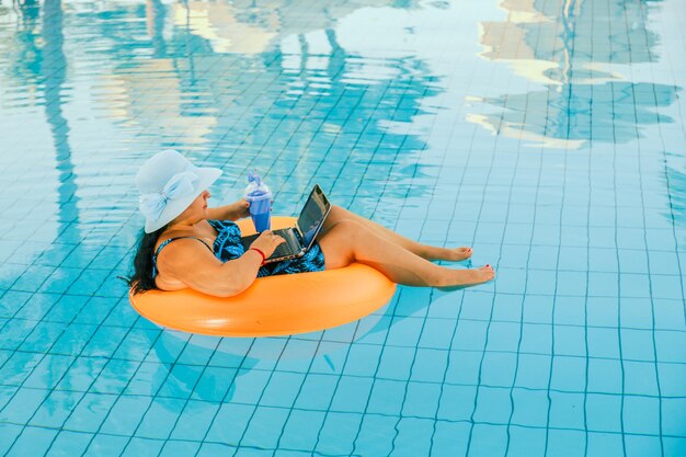 Mujer morena joven en un sombrero para el sol en la piscina en un círculo de natación con un cóctel y una computadora portátil