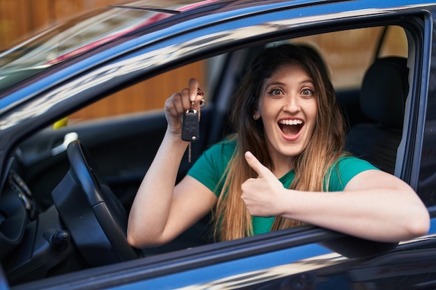 Mujer morena joven sosteniendo la llave del auto nuevo apuntando con el pulgar hacia el lado sonriendo feliz con la boca abierta