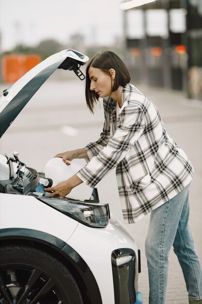 Mujer morena joven vertiendo un líquido en un motor en el capó del coche mientras está de pie en un estacionamiento. Mujer vistiendo chaqueta a cuadros, jeans