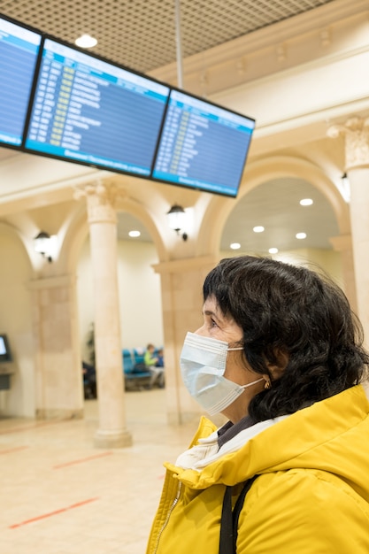 Mujer morena jubilado en una máscara protectora médica mira el tablero de vuelo en el aeropuerto