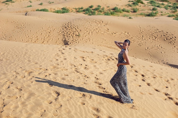 Foto una mujer morena con un largo vestido de leopardo se encuentra de espaldas en el desierto al atardecer, va a todas partes