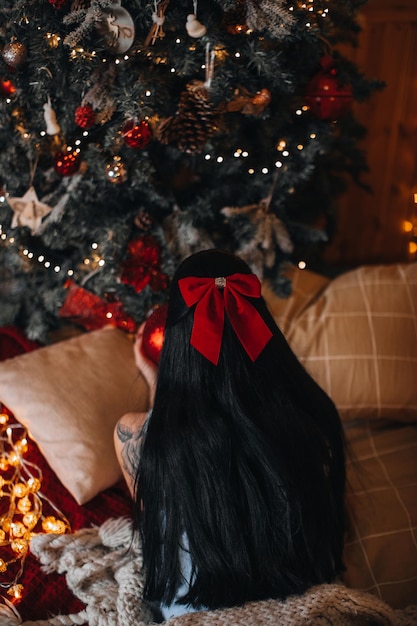Una mujer morena con un lazo rojo en el pelo cerca del árbol de Navidad con adornos festivos