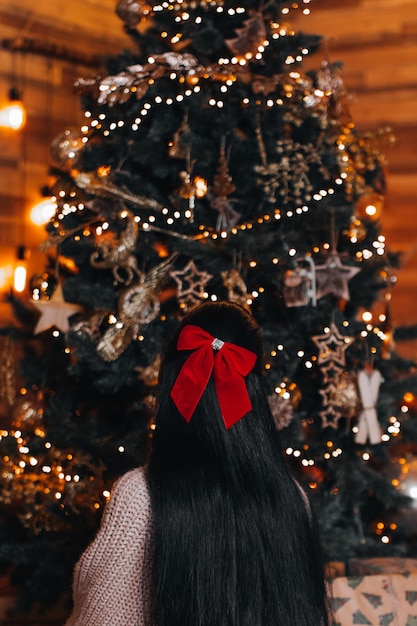 Una mujer morena con un lazo rojo en el pelo sentada cerca de un árbol de Navidad con adornos dorados