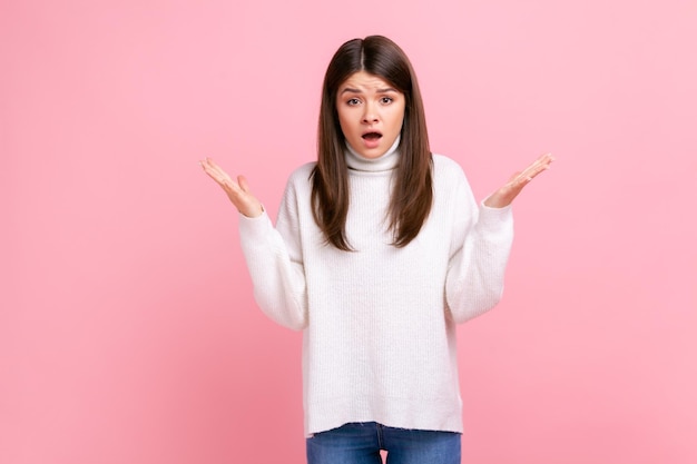 Mujer morena levantando las manos y mirando con expresión confusa peleando molesta por el conflicto usando un suéter blanco de estilo casual Foto de estudio interior aislada en fondo rosa