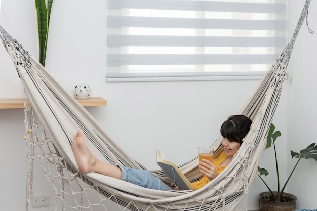 Mujer morena leyendo un libro y bebiendo jugo de fruta mientras se relaja en una hamaca cerca de la ventana