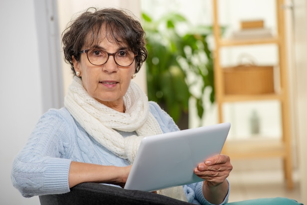 Mujer morena mayor encantadora con los vidrios usando la tableta digital en casa