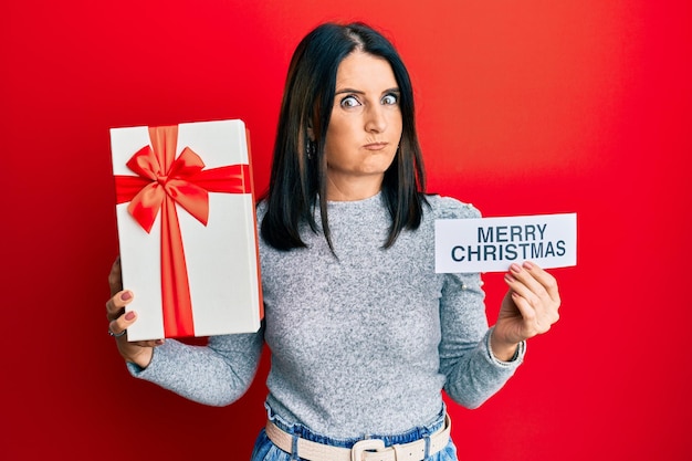 Mujer morena de mediana edad sosteniendo un mensaje de feliz navidad y regalando mejillas con cara graciosa. boca inflada con aire, atrapando aire.