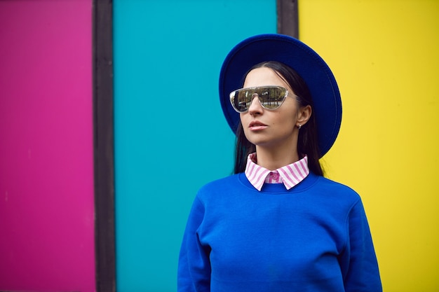 Foto mujer morena de moda en un gorro azul y un suéter y gafas de sol se encuentra en el contexto de las ventanas de colores en otoño