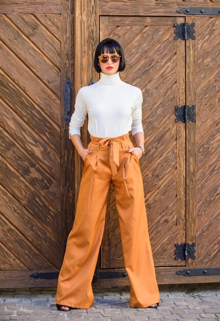 Mujer morena de moda stand al aire libre fondo de madera Chica con maquillaje posando en ropa de moda Traje de moda dama alta delgada Concepto de moda y estilo Mujer caminar en pantalones sueltos