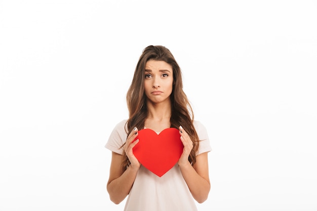 Mujer morena molesta en camiseta con corazón de papel sobre pared blanca