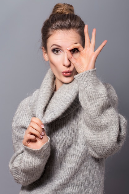 Mujer morena con moño peinado y maquillaje neutro posando en suéter gris sobre un fondo gris