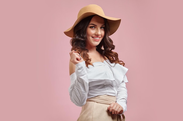 mujer morena muy sonriente con elegante sombrero beige y camisa blanca sobre rosa aislado