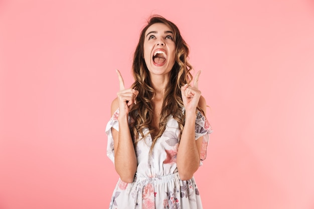 Mujer morena con el pelo largo vistiendo un vestido apuntando con el dedo hacia arriba, aislado en rosa