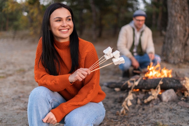 Mujer morena pensativa disfrutando de la recreación al aire libre mientras asaba malvaviscos sobre el fuego con su novio. Foto de stock