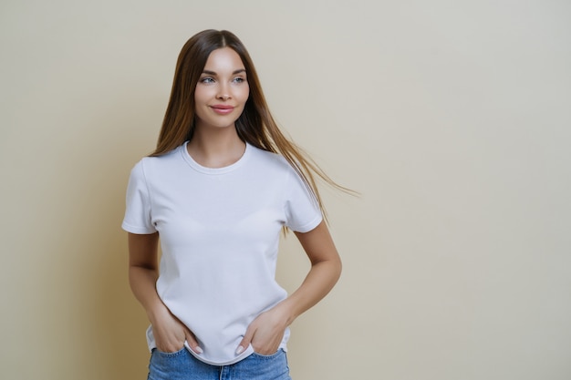 La mujer morena pensativa mira a un lado, usa una camiseta blanca informal, mantiene las manos en los bolsillos y mira