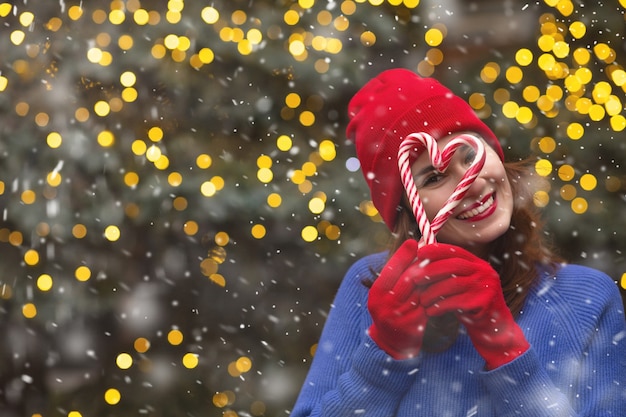 La mujer morena positiva viste un sombrero rojo y un suéter azul con caramelos cerca del árbol de Navidad durante las nevadas. Espacio para texto