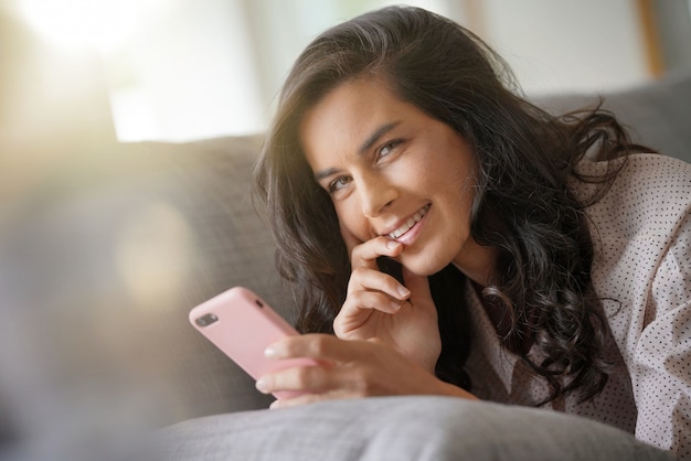 Mujer morena que se relaja en el sofá, conectado con el teléfono inteligente