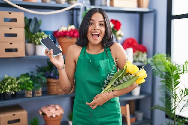 Mujer morena que trabaja en la floristería sosteniendo un teléfono inteligente celebrando loco y asombrado por el éxito con los ojos abiertos gritando emocionado.