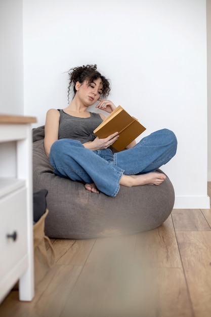 Foto mujer morena relajada acostada en la silla bolsa leyendo un interesante libro de papel disfrutando de fin de semana en casa