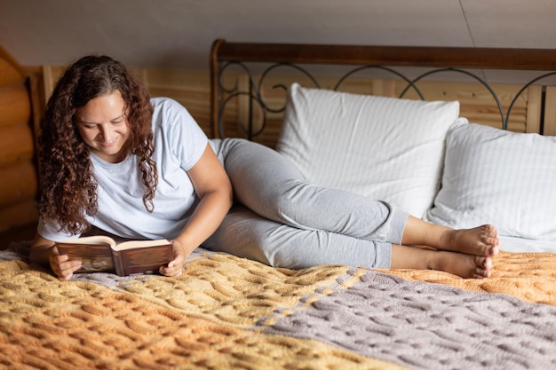 Mujer morena rizada adulta acostada en una cama doble cubierta con una colcha de felpa con un libro en las manos leyendo con cara feliz usando ropa de casa Tranquilo tiempo de ocio en casa