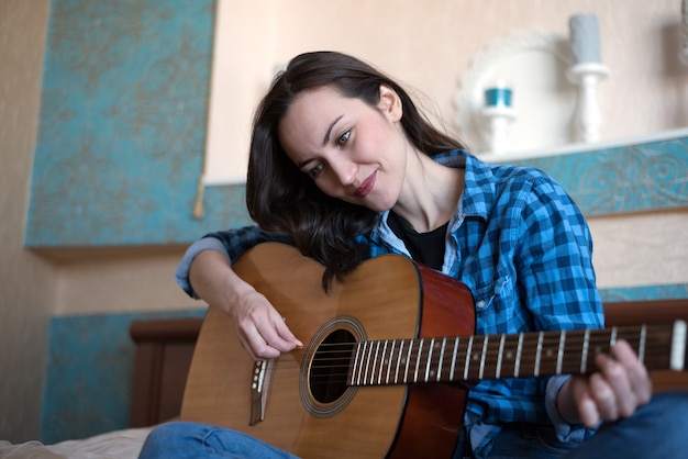Mujer morena sentada en la cama en el dormitorio con guitarra componiendo una canción - músico, compositor