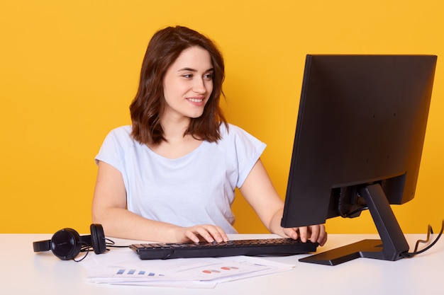 Mujer morena sentada en el escritorio blanco y escribe algo en el teclado mientras trabaja en línea, usando la computadora de la PC