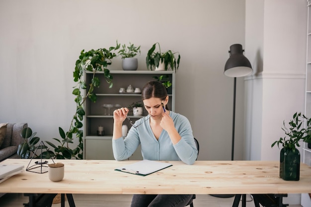 una mujer morena seria con una chaqueta azul está sentada en una mesa con papeles. Consulta empresarial